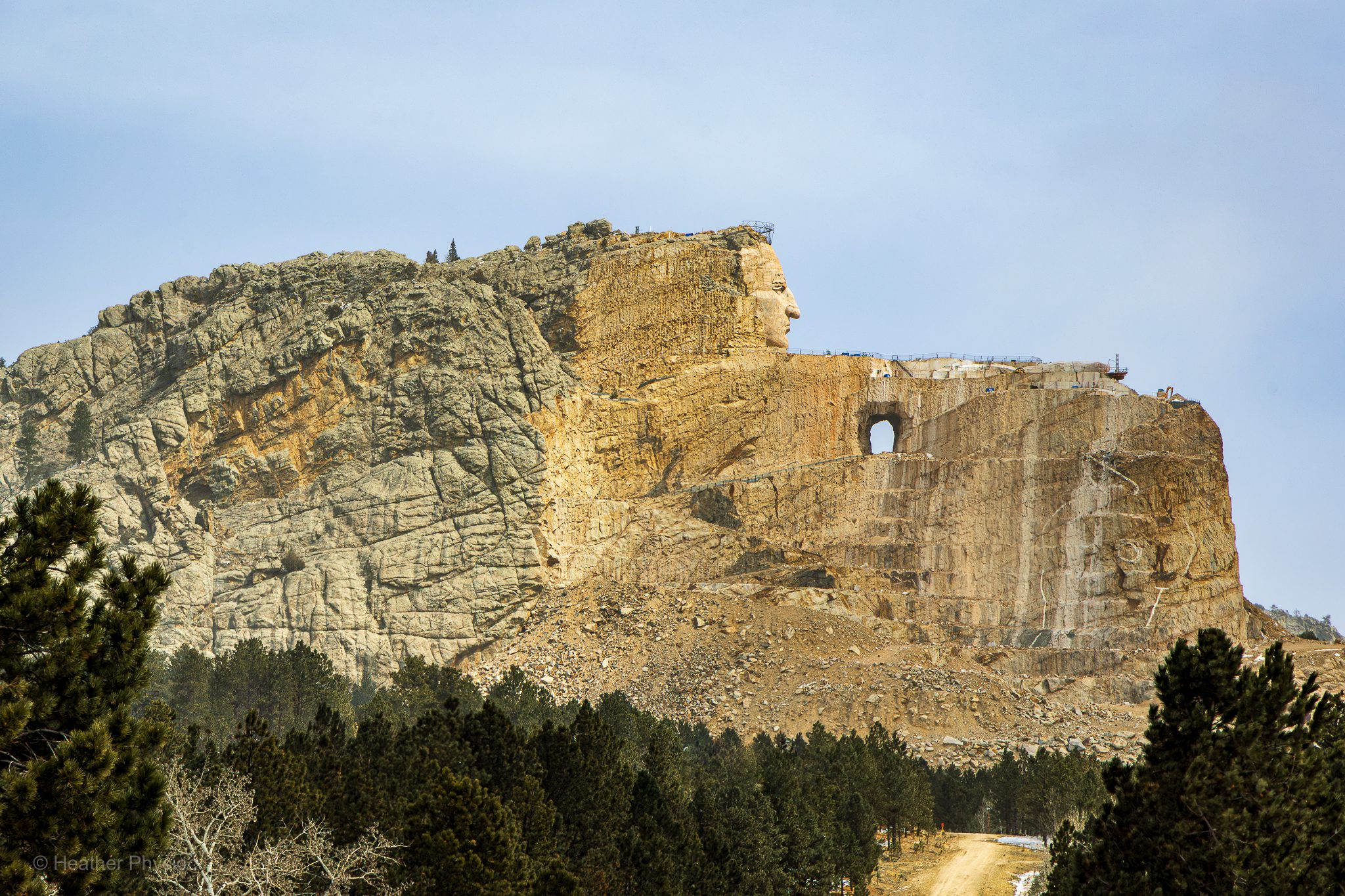 Wide view of the Crazy Horse Memorial under construction in South Dakota’s Black Hills, showing scaffolding and equipment around the completed head of Crazy Horse and the partially carved outline of his arm and horse emerging from the mountain above a forested foreground under a pale blue sky.