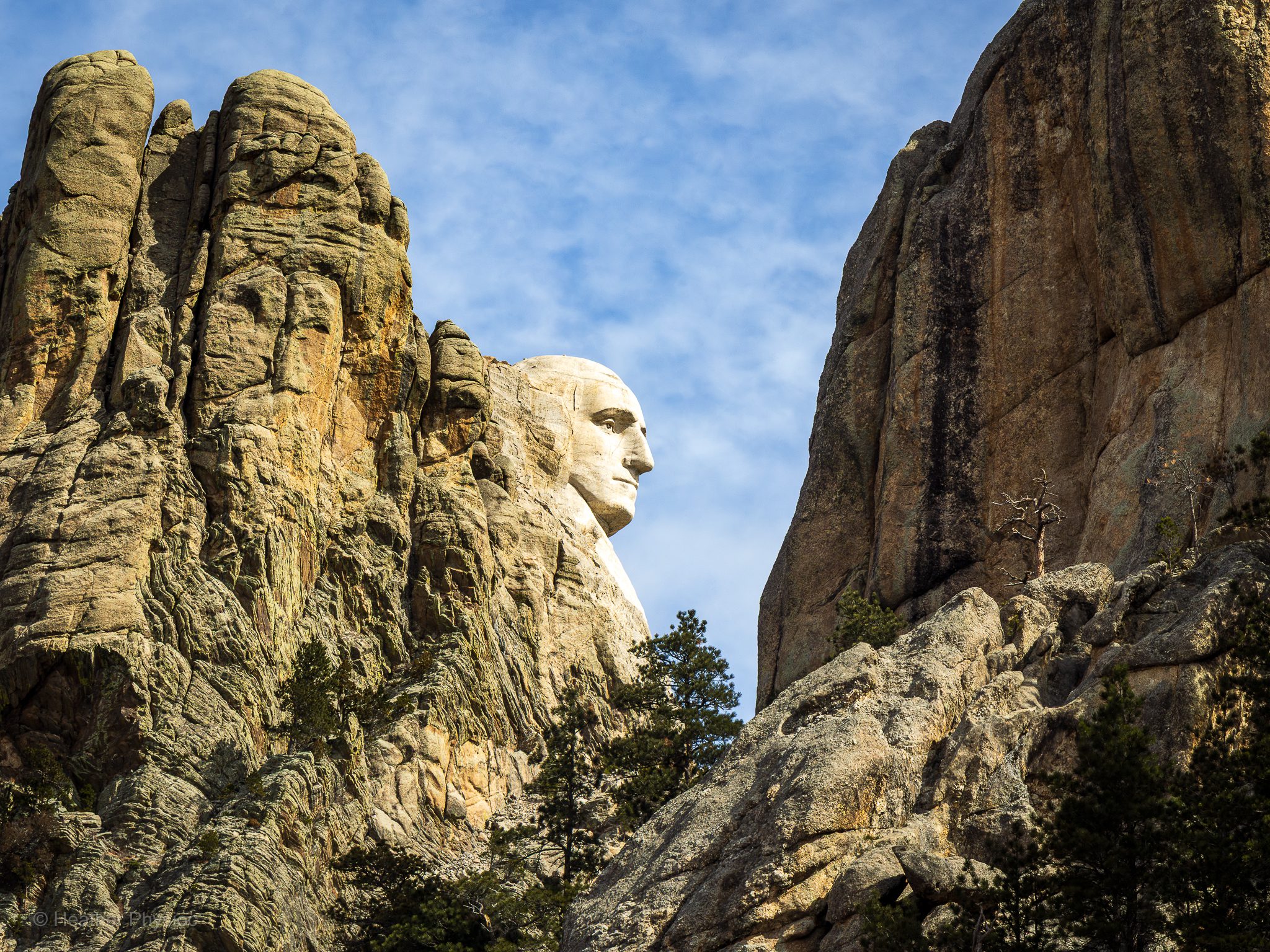 Side view of George Washington’s carved face on Mount Rushmore, framed dramatically between rugged granite cliffs and scattered pine trees under a partly cloudy blue sky in the Black Hills of South Dakota.