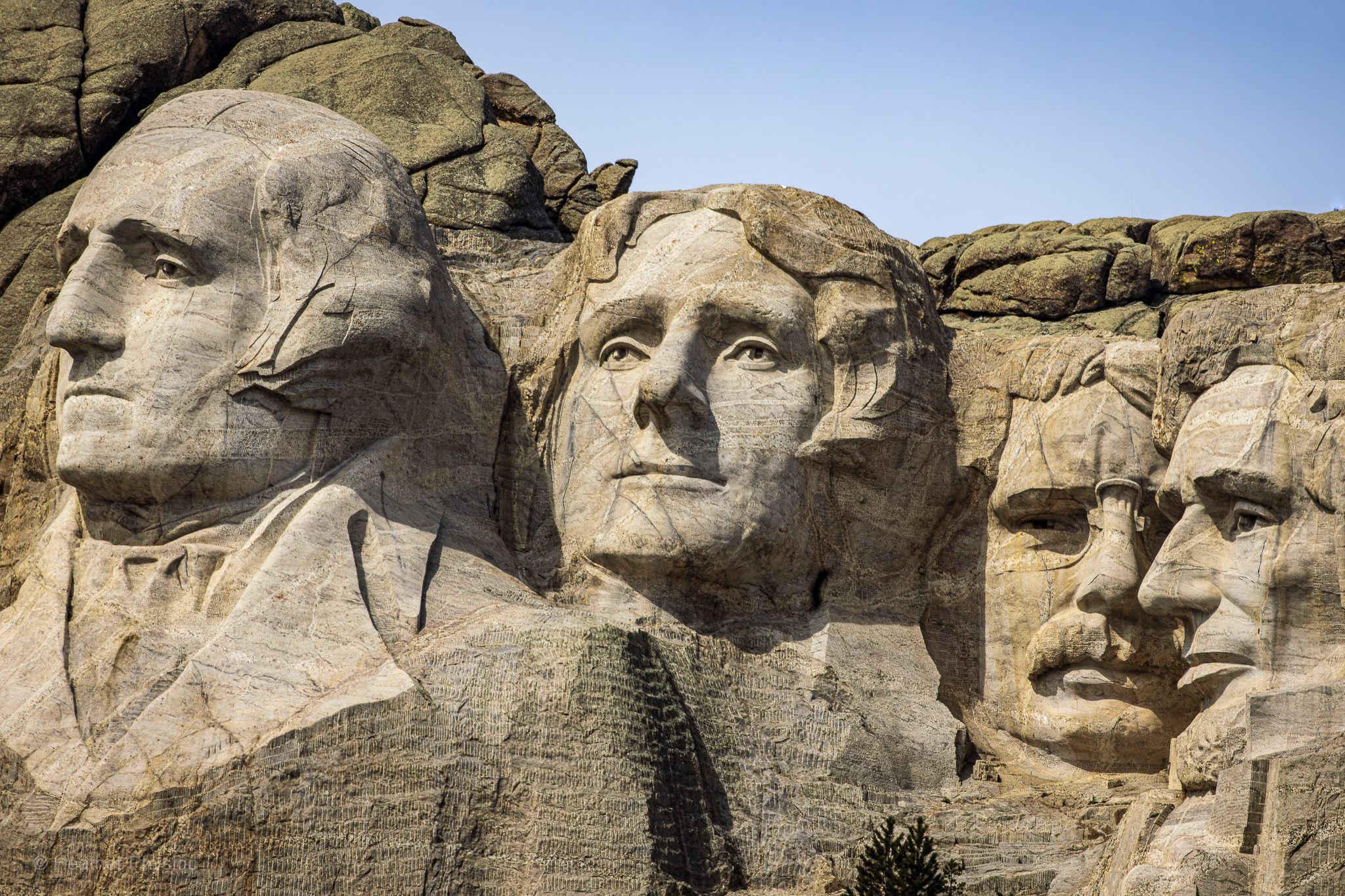 Close-up view of the Mount Rushmore National Memorial in South Dakota, showing the colossal granite faces of four U.S. presidents—George Washington, Thomas Jefferson, Theodore Roosevelt, and Abraham Lincoln—carved into the mountainside against a clear blue sky.