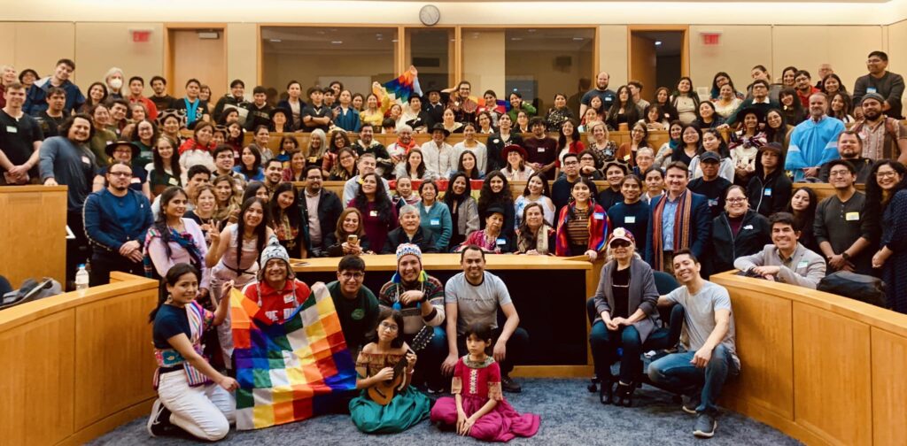 Community educational event at Harvard University for the 2023 Quechua Alliance annual meeting: A large group of people fills a lecture hall at Harvard University during the 2023 Quechua Alliance annual meeting. Participants of diverse ages and backgrounds smile toward the camera, some wearing traditional Andean clothing and woven textiles, while others hold colorful Wiphala flags representing Indigenous unity. At the center of the gathering, Dr. Américo Mendoza-Mori stands among community members, educators, and students, celebrating Quechua language and cultural education in an atmosphere of pride and solidarity.