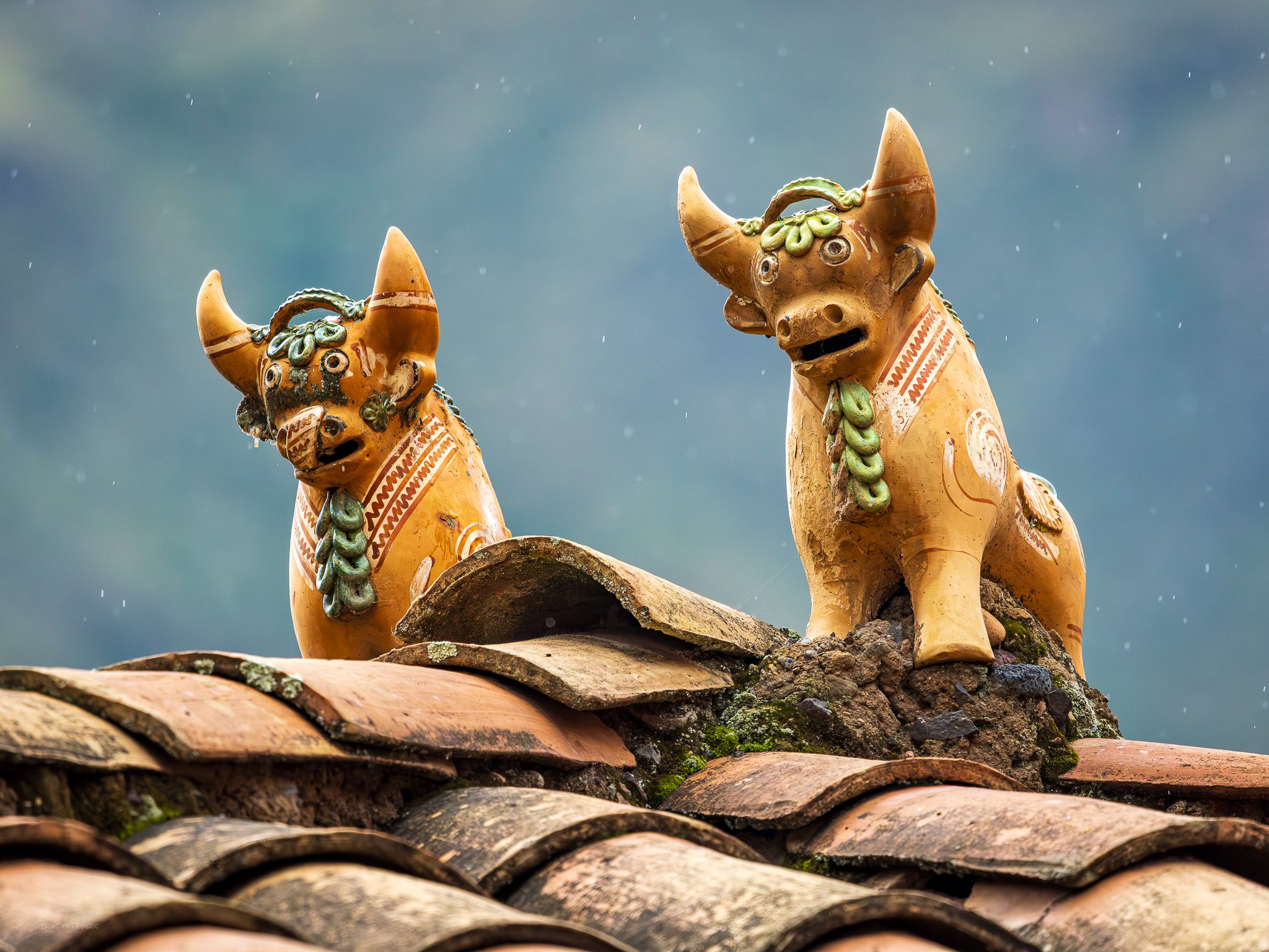 Pucara bulls serve as iconic protectors atop Cusco's red-tiled roofs: Two handcrafted ceramic Pucara bulls stand side by side on a red-tiled roof, their ochre bodies adorned with green decorative details and patterned carvings. Set against a misty Andean mountain backdrop, the sculptures’ expressive faces and upturned horns symbolize protection, prosperity, and strength in Quechua culture. A few raindrops fall softly, adding a sense of quiet reverence to this enduring Andean tradition.