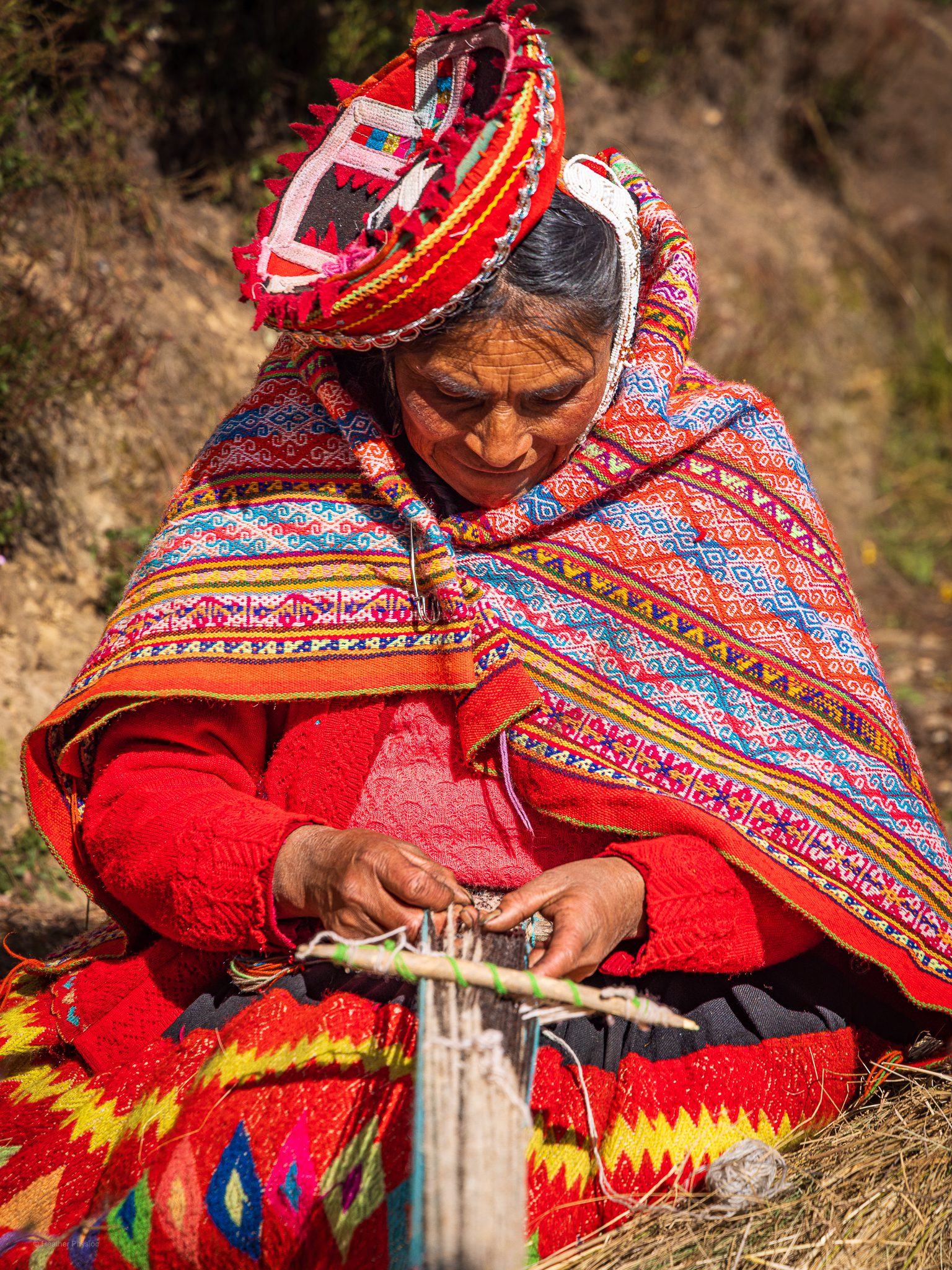 Quechua abuela hand-weaves alpaca wool using traditional methods: An elderly Quechua woman sits outdoors weaving on a traditional backstrap loom, her focus steady on the intricate threads between her hands. She wears a vivid red sweater and a handwoven shawl decorated with detailed geometric patterns in bright pink, orange, blue, and yellow. A traditional round hat adorned with ribboned edges rests on her head, tilted by the sun. The golden light highlights the textures of her clothing and the craftsmanship of her weaving—an art form passed down through generations of Andean women.
