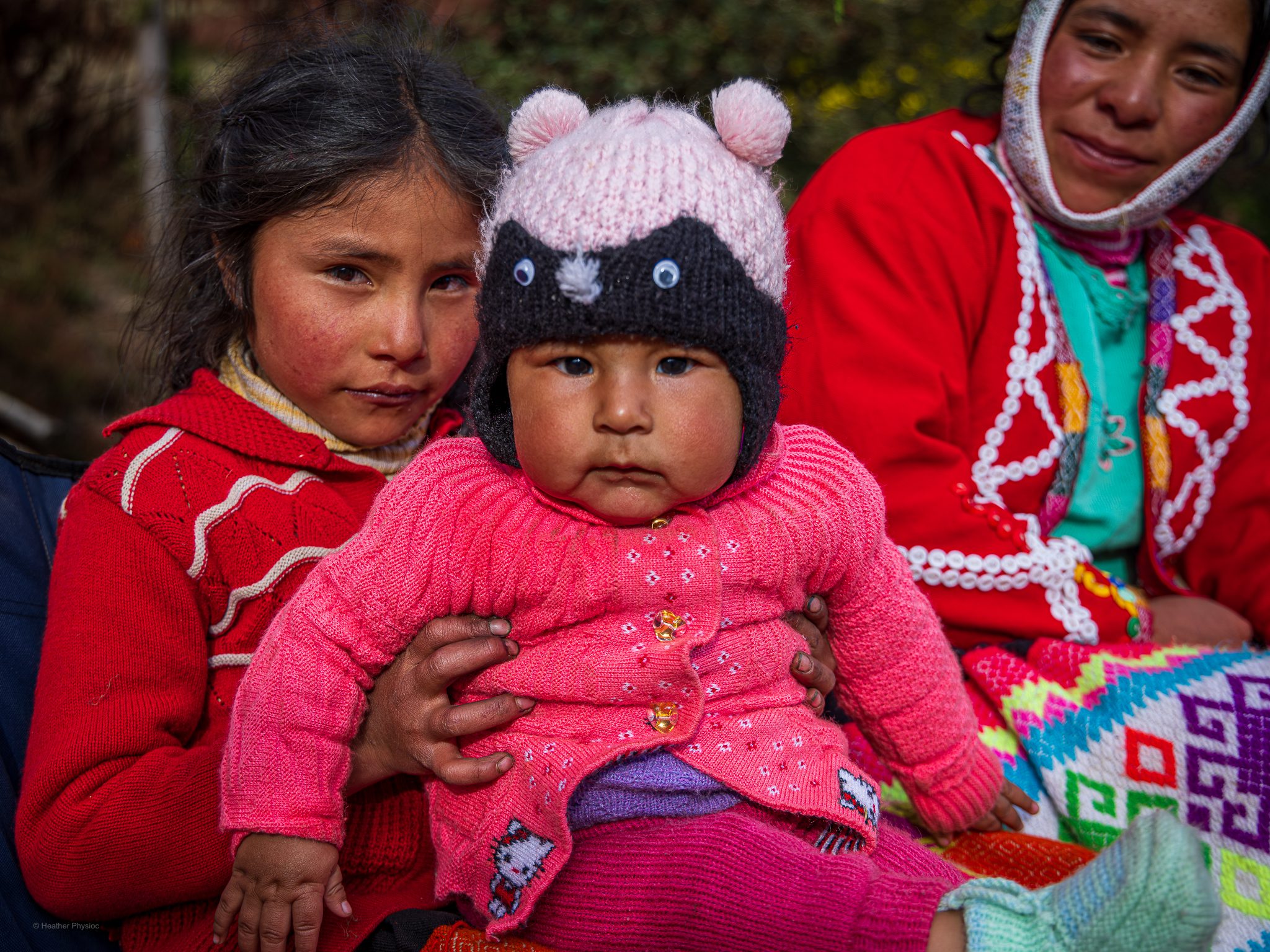 A young Quechua girl wearing a red sweater holds a bundled baby dressed in pink knit clothing and a hat with animal ears and button eyes. Both sit outside in soft natural light, their cheeks flushed from the mountain air. An older woman in the background, also in traditional colorful Andean clothing, watches with a gentle smile. The image captures the warmth and shared caregiving that are central to family life in Quechua communities.