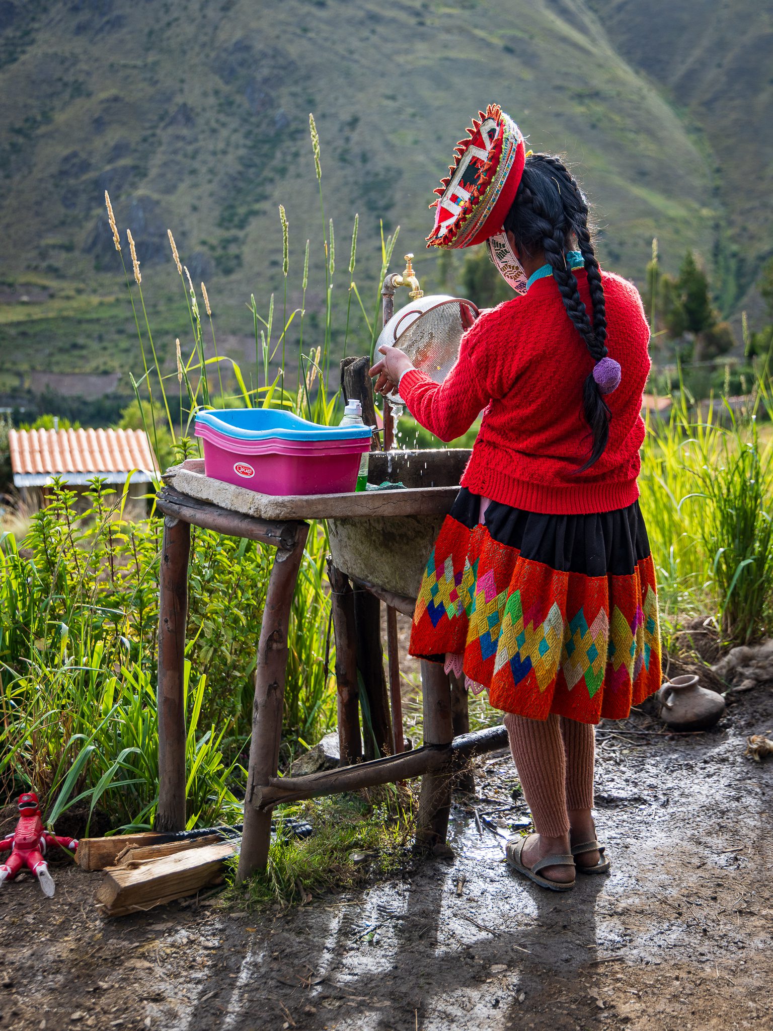 Young Quechua woman washes dishes in the shadows of the Andes: A young Quechua girl, wearing a traditional red sweater, black skirt with bright geometric patterns, and a round embroidered hat, stands at an outdoor sink washing dishes. Water trickles from a brass faucet as she cleans a metal bowl beside a pink plastic basin. Her long braids fall down her back as late afternoon light illuminates the green mountain slopes surrounding her rural home near Patacancha in the Peruvian Andes.