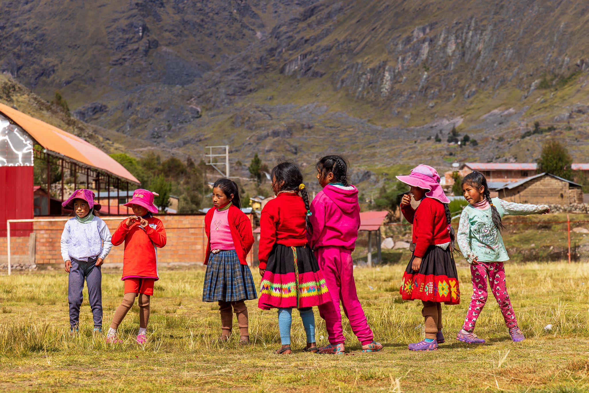 A group of eight Quechua schoolgirls stands together on a grassy field in the Andean highlands, playing an outdoor game under the mountain sun. They wear bright clothing — red sweaters, colorful skirts, pink hats, leggings, and sneakers — reflecting both traditional Andean patterns and modern styles. A few look at one another mid-conversation, while others glance toward the camera. Behind them are rustic brick school buildings and a corrugated-roof structure, with rocky mountain slopes rising in the background.