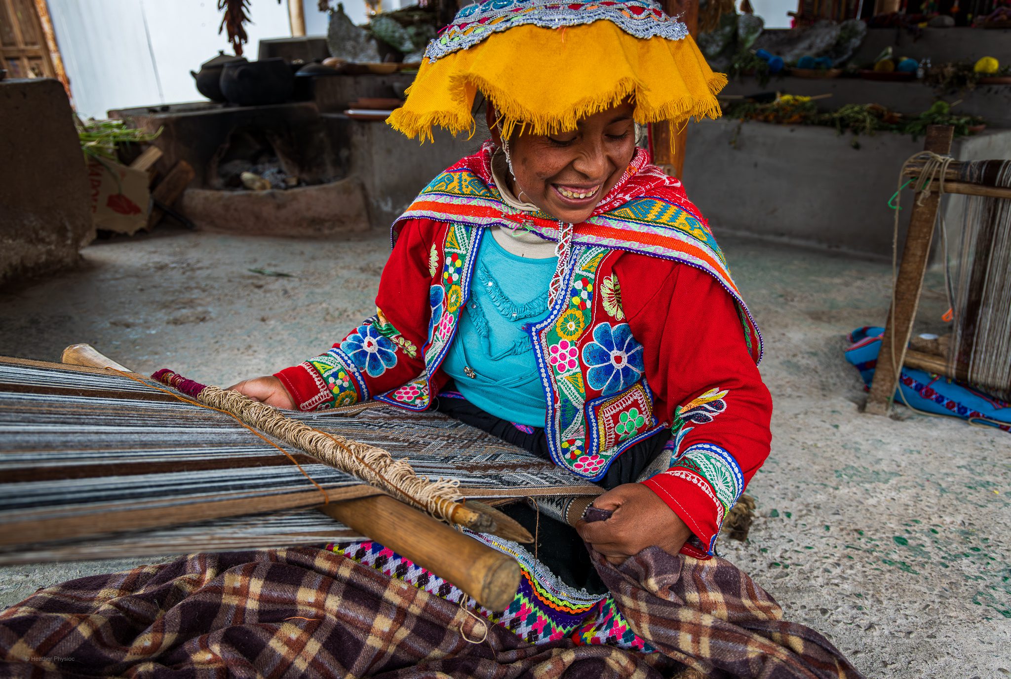 A Quechua woman smiles as she weaves alpaca wool on a traditional backstrap loom in Cusco. She wears a vividly embroidered red jacket decorated with floral designs, a turquoise blouse, and a yellow fringed hat. Her hands skillfully guide the threads, creating a patterned textile stretched between wooden rods. The soft light highlights the colorful yarns and intricate craftsmanship, reflecting the deep Andean weaving traditions passed down through generations.