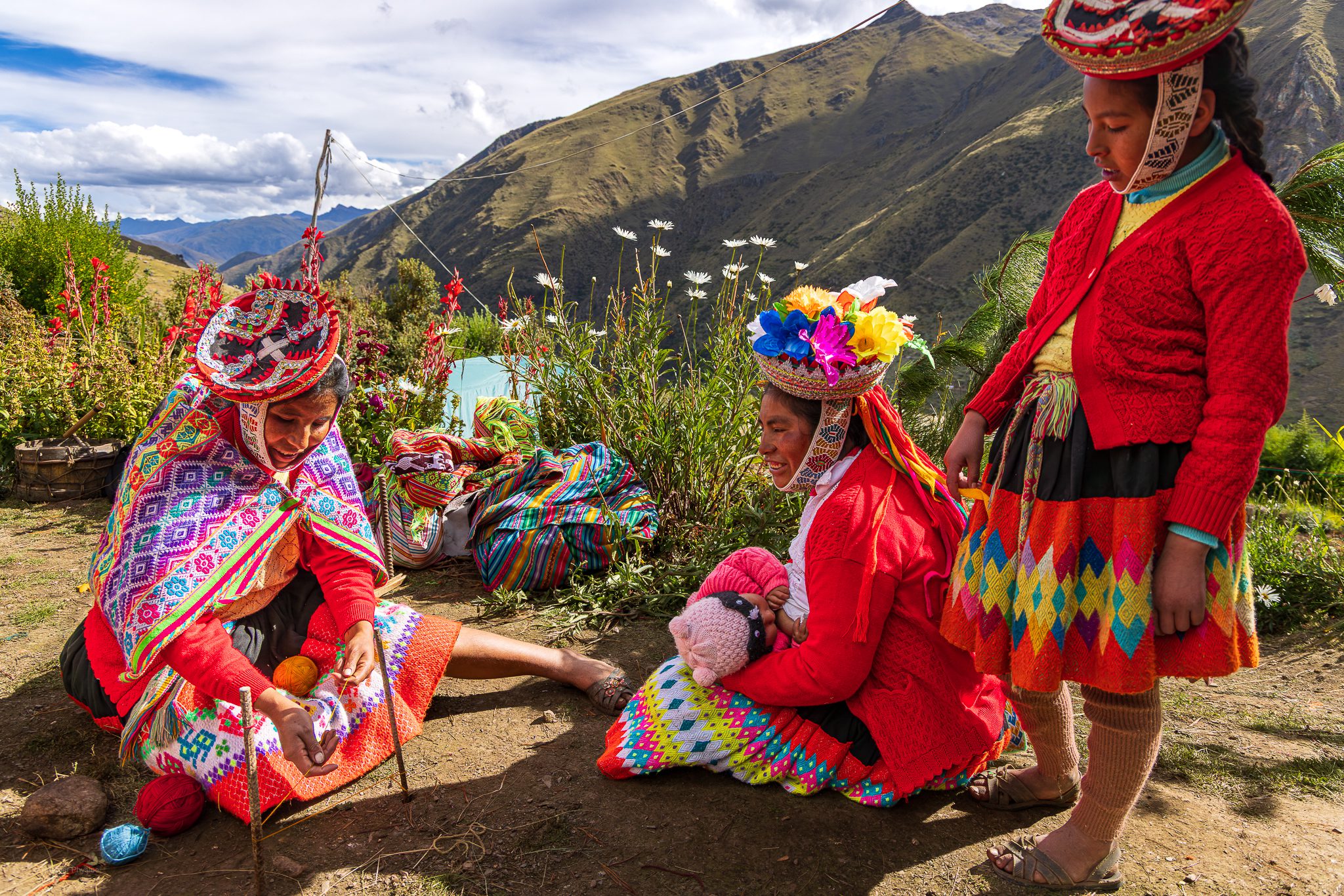 Three Quechua women, one a young teenager and two adult women weaving, with a young baby breastfeeding. All are dressed in brightly dyed wools of red, pink, yellow and purple, from their alpaca weaving. Three Quechua women and girls sit and stand outdoors in the Andean highlands, surrounded by blooming flowers and mountain views. They wear traditional handwoven clothing—bright red sweaters, patterned skirts, and elaborately embroidered hats adorned with ribbons and flowers. One woman sits cross-legged on the ground spinning colorful wool, while another cradles a baby dressed in pink. The warm sunlight highlights the vivid textiles and the women’s deep connection to their heritage and landscape.
