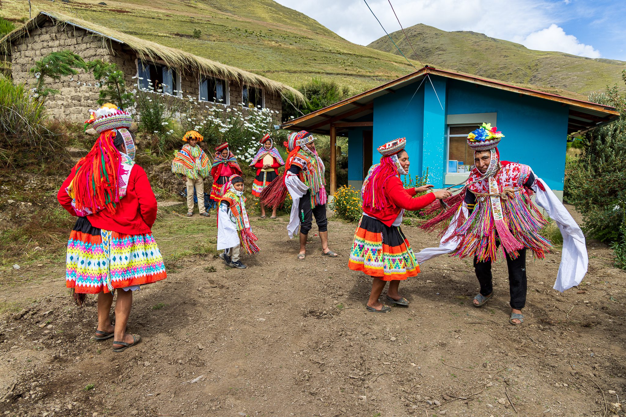 Quechua Welcome Dance: A lively outdoor scene in a rural Andean village shows men, women, and children dressed in bright traditional garments dancing and laughing near adobe houses with thatched roofs and a turquoise-painted building. The participants wear layered skirts, woven belts, and headdresses adorned with multicolored tassels and ribbons. Mountains rise in the background under a partly cloudy sky, creating a joyful, celebratory atmosphere.