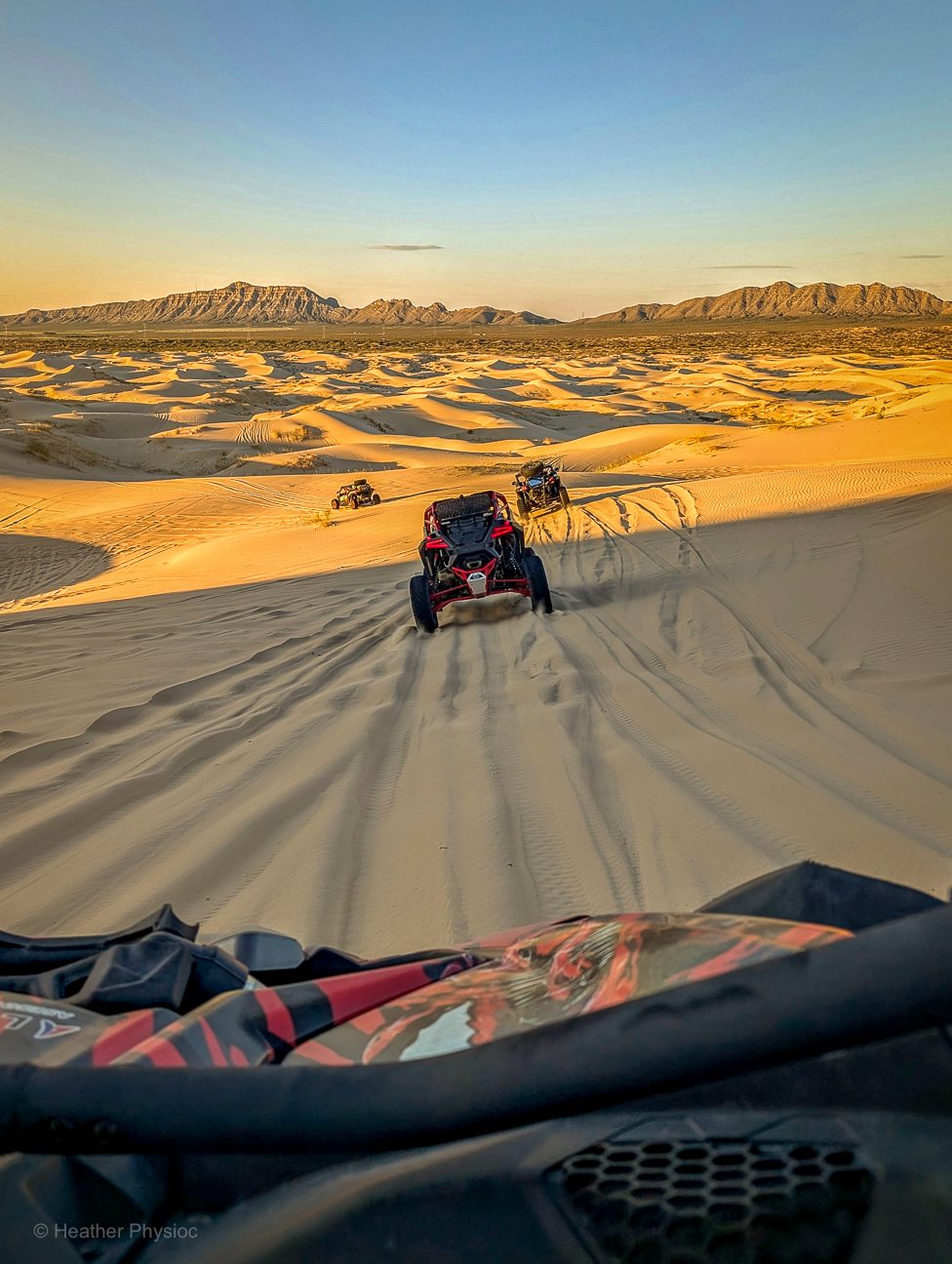 All-terrain dune buggies rumble across a vast desert of rolling golden sand, their deep tire tracks carving fresh lines through the rippled dunes. The lead buggy in vivid red dominates the foreground, while others trail behind under a warm amber sky. In the distance, a rugged mountain range rises against the horizon, contrasting with the sweeping, endless expanse of sand. The image captures both exhilaration and immensity.
