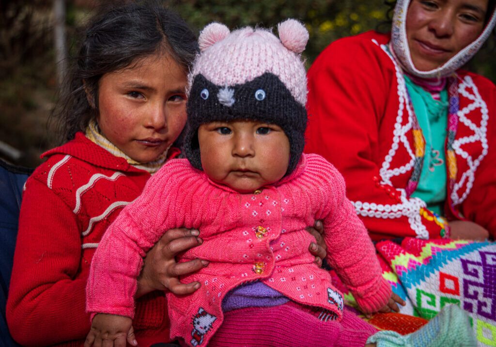 A young Quechua girl wearing a red sweater holds a bundled baby dressed in pink knit clothing and a hat with animal ears and button eyes. Both sit outside in soft natural light, their cheeks flushed from the mountain air. An older woman in the background, also in traditional colorful Andean clothing, watches with a gentle smile. The image captures the warmth and shared caregiving that are central to family life in Quechua communities.